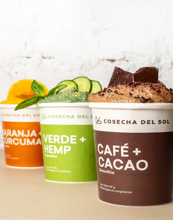 Tres empaques de smoothie, Naranja + Cúrcuma, Verde + Hemp y Café + Cacao, frente a una pared blanca.