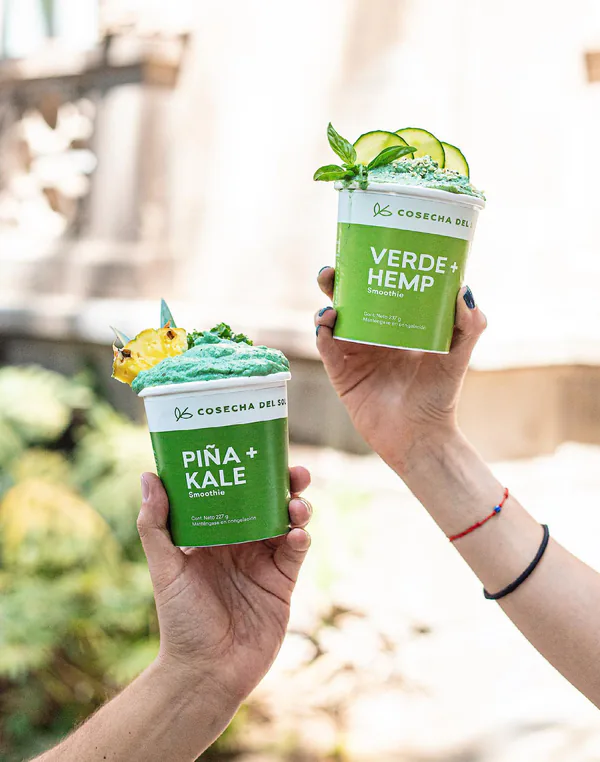 Dos manos levantan empaques de Piña + Kale y Verde + Hemp con el smoothie servido adentro.