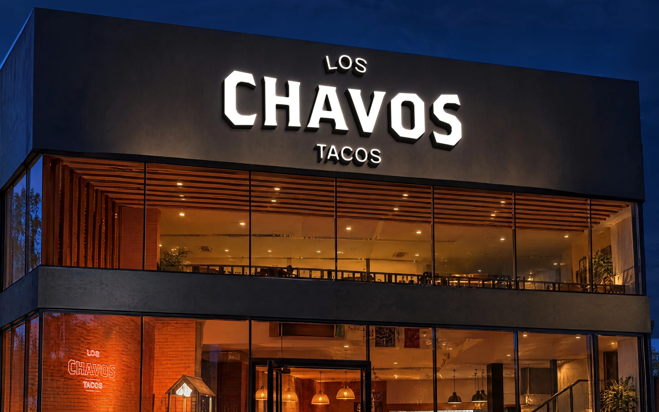 Fachada de Los Chavos de noche: arriba, el letrero con el logotipo prendido en blanco; abajo, dos pisos de ventanales con luz cálida.