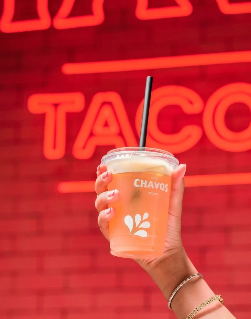 Vaso con el logotipo de Los Chavos en una mano, frente a un letrero de neón rojo.