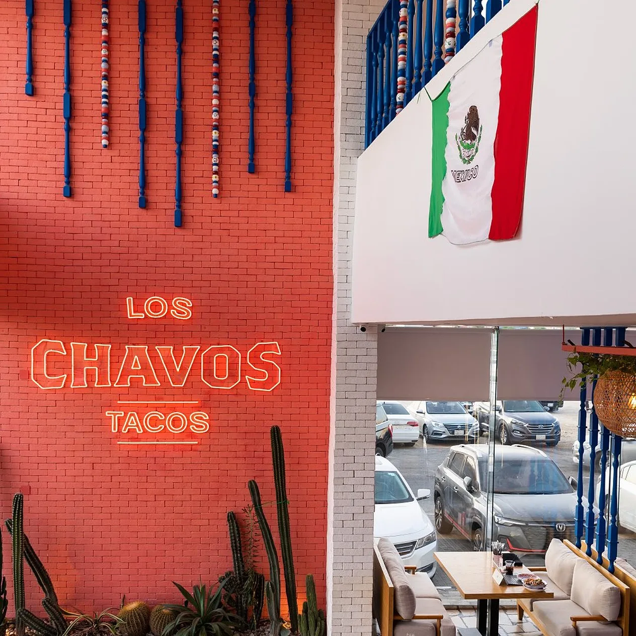 El neón rojo de Los Chavos en un muro de ladrillo con cactus al pie y, a la derecha, el salón, con la bandera de México arriba y el estacionamiento detrás.